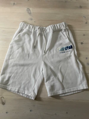 Vita Trapstar shorts med brodyr - Snygga vita shorts från Trapstar med broderad logga i blå och vit på ena benet. Shortsen har snörning i midjan och är gjorda i mjuk bomull, perfekta för sommardagar. Avslappnad passform och sidofickor för en chill look.