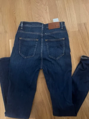 Mörkblå skinny jeans från tiger of sweden - Snygga mörkblå skinny jeans från Ivy med klassiska fem fickor och bruna detaljer vid bakfickorna. Jeansen har en smal passform och är tillverkade i stretchigt denim för extra komfort. Perfekta för en trendig och avslappnad look.