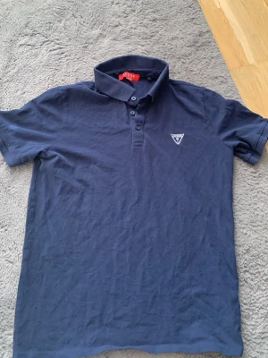 Mörkblå pikétröja från Guess - Snygg mörkblå pikétröja från Guess med klassisk krage och knappslå framtill. Tröjan har korta ärmar och en liten logotyp på bröstet. Perfekt för en stilren och avslappnad look.