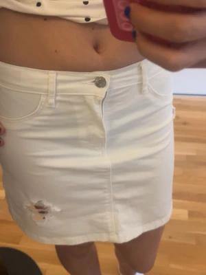 Vit jeanskjol med slitning - Snygg vit kort jeanskjol med klassiska fickor fram och bak samt bälteshällor. Kjolen har en cool slitning framtill som ger en trendig look. Perfekt att matcha med en crop top eller t-shirt för en avslappnad stil.