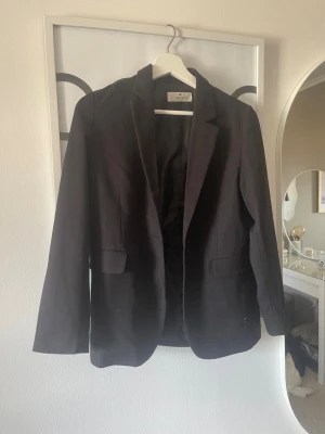 Svart kavaj från H&M - Stilren svart kavaj från H&M med klassisk passform och två framfickor med lock. Kavajen har enkelknäppning och är helfodrad, perfekt för en clean och snygg look. Materialet är mjukt och följsamt, vilket gör den bekväm att bära hela dagen.