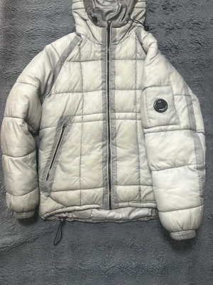 Grå pufferjacka från C.P. Company - Säljer en grå pufferjacka från C.P. Company med huva och dragkedja framtill. Jackan har quiltad design, två snedställda fickor med dragkedja och en ikonisk linsdetalj på ärmen. Perfekt för kalla vinterdagar och riktigt snygg att bära.