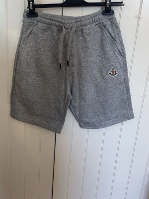 Grå Moncler shorts med snörning - Snygga grå shorts från Moncler med elastisk midja och snörning. Mjukt material och två sidofickor. Moncler-logga på vänster ben ger en exklusiv touch. Perfekta för en avslappnad och sportig stil.