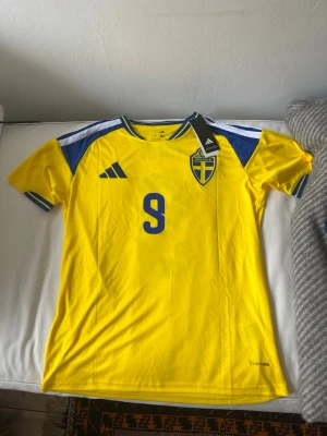 Sverige fotbollströja Isak #9 Adidas - Gul och blå Sverige fotbollströja från Adidas med nummer 9 och namnet Isak på ryggen. Tröjan har korta ärmar, blå detaljer på axlarna och det svenska landslagets emblem på bröstet. Tillverkad i lätt och ventilerande material. Säljer pågrund av att jag vill ha ett annat namn på ryggen. (Helt oanvänd)