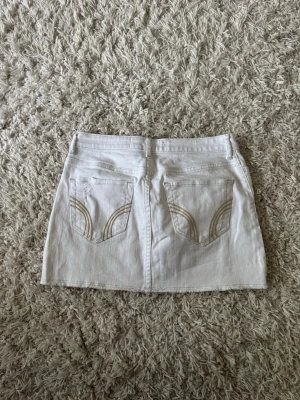 Vit kort jeanskjol från Hollister - Vit jeanskjol från Hollister med klassiska fem fickor och snygga broderade detaljer på bakfickorna. Kjolen är kort och har en enkel, stilren design som passar perfekt till sommarens outfits.