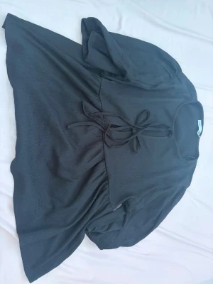 Svart blus med knytband - Svart blus med rund halsringning och knytband framtill. Blusen har lätt puffade långärmar och är tillverkad i ett mjukt, tunt tyg med diskret struktur. Passformen är avslappnad och modellen är något längre baktill.
