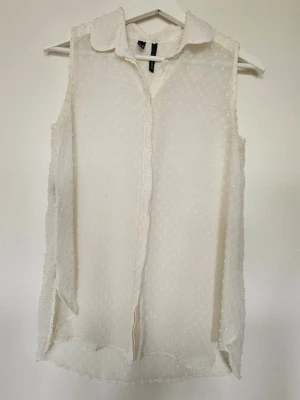 Vit ärmlös blus från Mango  - Säljer en vit ärmlös blus från Mango med diskret prickigt mönster och klassisk krage. Blusen är gjord i ett tunt och lätt material som ger en luftig känsla. Perfekt för att skapa en stilren och fräsch look.