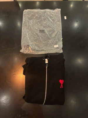 Svart hoodie med dragkedja från Ami - Svart hoodie från Ami med dragkedja framtill och huva med dragsko. På bröstet finns Ami:s ikoniska röda hjärta med bokstaven A. Hoodien har ribbade muddar och fickor framtill. Perfekt för en stilren och trendig look.