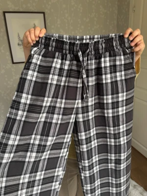 Rutiga pyjamasbyxor i svart och vitt - Mysiga pyjamasbyxor med svartvitt rutmönster och dragsko i midjan. Byxorna har en avslappnad passform och elastisk midja för extra komfort. Perfekta för chillkvällar hemma eller när du vill vara extra bekväm.