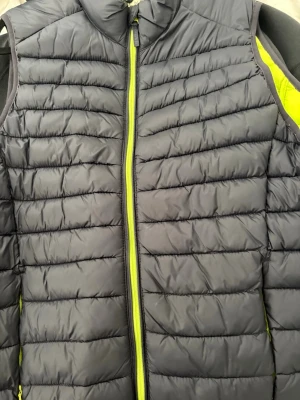 Svart dunväst från Blend med neonfoder - Snygg svart dunväst från Blend med quiltad design och dragkedja i neonfärg. Insidan har ett coolt neongult foder som ger västen en extra edge. Perfekt för lager-på-lager och streetstyle. Materialet är lätt och smidigt, vilket gör den enkel att bära.