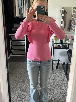 Rosa topp med volangdetaljer - Snygg rosa långärmad topp med volanger längs axlarna. Modellen är figurnära och toppen har en rund halsringning. Perfekt för dig som vill sticka ut med en färgglad och trendig look.