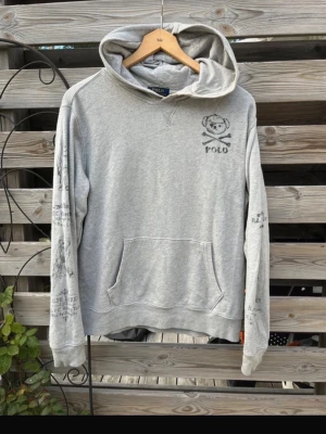 Grå hoodie från Polo Ralph Lauren🙌🤩 - Grå hoodie från Polo Ralph Lauren med tryck av björn och text på bröstet samt detaljerade tryck längs ärmarna. Klassisk känguruficka framtill och huva. Perfekt för en avslappnad och stilren look.🤩