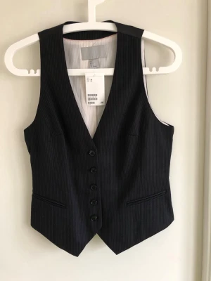Svart randig väst från H&M - Stilren svart väst med tunna vita ränder från H&M. Västen har djup v-ringning, fem knappar framtill och två fickor. Baksidan är i slätt tyg med justerbart spänne. Perfekt för en dressad och trendig look.