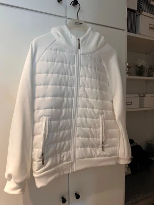 Vit pufferjacka med huva - Snygg vit pufferjacka med quiltade paneler och dragkedja framtill. Jackan har två fickor med dragkedja och en mysig huva. Tillverkad i ett mjukt material som håller värmen under kalla dagar. Perfekt för dig som vill ha en stilren och enkel look.