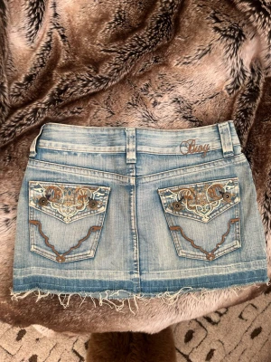 Ljusblå jeanskjol från Siný med brodyr - Cool ljusblå jeanskjol från Siný med slitna kanter och snygg brodyr på bakfickorna. Kjolen har klassisk femficksdesign, bälteshällor och detaljer i brunt och vitt. Perfekt för dig som gillar en avslappnad och trendig stil.
