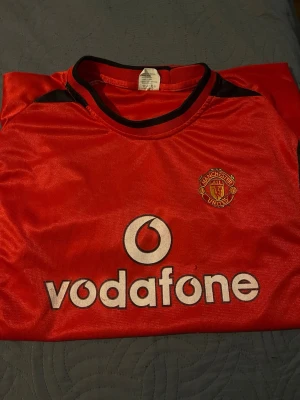 Manchester United tröja V. Nistelrooy - Klassisk röd Manchester United matchtröja med svart detalj, Vodafone-logga på framsidan och klubbmärke på bröstet. På ryggen står det V. Nistelrooy och nummer 10. Perfekt för fotbollsfans! Materialet är polyester.