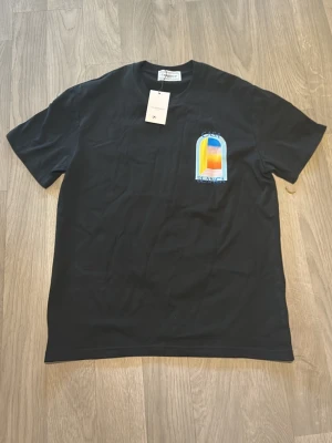 Svart t-shirt från casablanca med tryck - Svart t-shirt från casablanca med färgglatt tryck på bröstet i blått, orange och gult. Klassisk passform med rund hals och korta ärmar. Perfekt för en avslappnad och trendig stil.