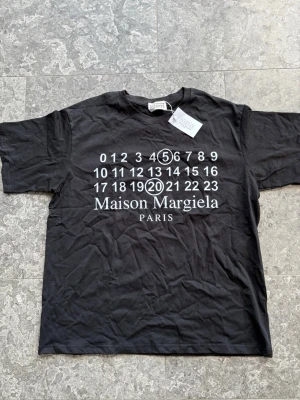 Maison Margiela svart t-shirt - Svart t-shirt från Maison Margiela med ikoniskt tryck av siffror och logga på bröstet. Klassisk rund hals och korta ärmar. Tillverkad i mjuk bomull som känns skön mot huden. Perfekt för dig som gillar streetstyle och designerplagg.