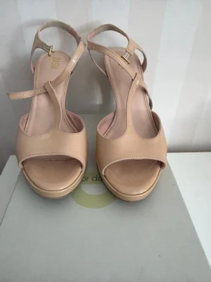 Beige högklackade pumps med remmar - Snygga beige pumps från Vagabond med hög klack och öppen tå. Skorna har eleganta T-remmar och justerbart spänne vid ankeln. Tillverkade i glansigt skinnliknande material som ger en stilren look. Perfekta för att lyfta vilken outfit som helst.