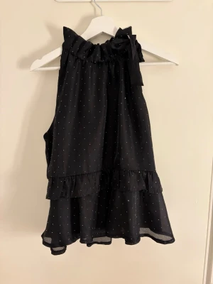 Svart ärmlös blus med volang - Snygg svart blus med små stenar, volangkant nedtill och runt halsen samt en söt rosett vid kragen. Blusen är ärmlös och har en luftig, loose passform som ger en avslappnad men chic look.