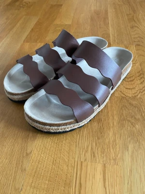 Bruna tofflor från Esprit - Trendiga tofflor från Esprit med tre breda vågformade remmar i brunt läder. Tofflor är knappt använda.
