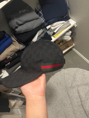 Äkta svart gucci keps svart färg storlek M - Säljer en svart keps från Gucci med det klassiska GG-monogrammönstret över hela kepsen. På sidan finns en grön och röd rand som ger den en ikonisk look. Kepsen har böjd skärm och justerbar rem bak. Perfekt för dig som vill ha en stilren och lyxig accessoar.