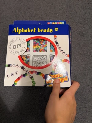 Alphabet beads DIY-kit - DIY-kit med färgglada plastpärlor och vita bokstavspärlor från Playbox of Sweden. Perfekt för att skapa personliga smycken som armband och halsband med egna texter och budskap. Innehåller pärlor i olika former och färger för kreativt pyssel.