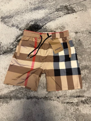 Rutig Burberry shorts med snörning - Säljer ett par klassiska Burberry shorts med det ikoniska rutiga mönstret i beige, svart, vit och röd. Shortsen har elastisk midja med snörning och en bakficka. Perfekta för sommaren och riktigt snygga till streetwear-stilen.