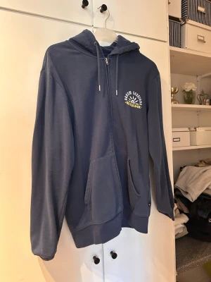 Blå zip hoodie från Quiksilver - Mörkblå hoodie med dragkedja från Quiksilver. Stor tryckt logga i vitt och gult på ryggen och mindre tryck på bröstet. Utrustad med huva och dragsnören samt en stor magficka. Perfekt för en avslappnad streetwear-look.