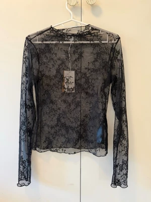 Svart långärmad spetstopp - Snygg svart långärmad topp i transparent spets med blommigt mönster. Tunn och figurnära modell som passar perfekt till lager-på-lager. Toppen har rund halsringning och är tillverkad i ett lätt och stretchigt material.