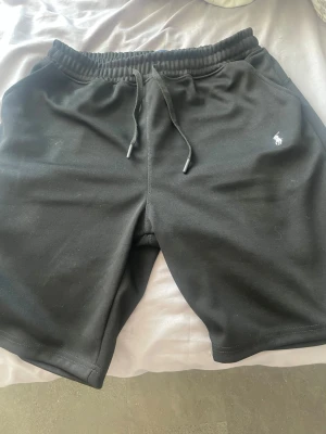 Svarta shorts från Polo Ralph Lauren - Säljer ett par svarta shorts från Polo Ralph Lauren med dragsko i midjan och fickor på sidorna. Klassisk logga broderad i vitt på ena benet. Sköna och stilrena shorts i mjukt material, perfekta för sommaren. Storlek XL hade sagt att den också passar L