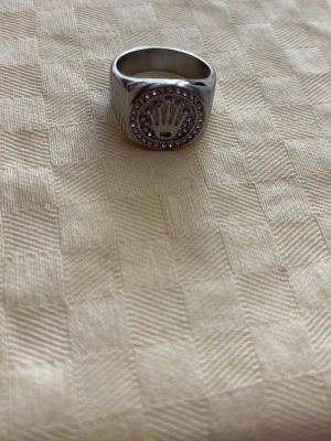 Silverfärgad ring med stenar - Snygg silverfärgad ring med rund front och gnistrande stenar runt en krona. Ringen har en bred design och är tillverkad i metall med blanka ytor. Perfekt för dig som gillar statement-smycken och vill sticka ut.