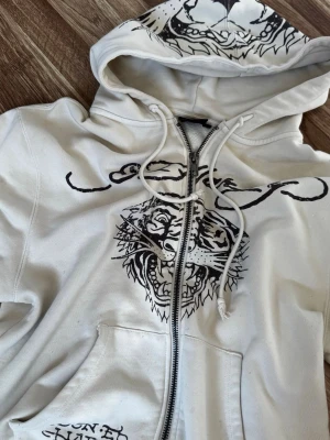 Vit Ed Hardy x Urban Outfitters hoodie med tigertryck - Säljer en vit hoodie från Ed Hardy x Urban Outfitters med dragkedja, huva, fickor och snörning. Stort svart tigertryck framtill och Ed Hardy-logga över bröstet. Huvan och armarna har också tryck. Perfekt för dig som gillar streetstyle och unika prints. Väldigt fint skick syns knappt att den är använd ett antal gånger! Priset är diskuterbart vid snabbt köp!