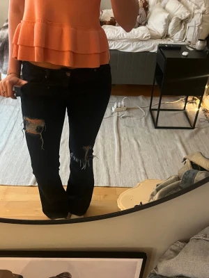 Mörkblåa slitna jeans med bootcut - Säljer ett par mörkblåa jeans med bootcut och stora slitningar på båda knäna. Jeansen har klassisk femficksdesign och låg midja. Perfekta för en avslappnad och trendig look.