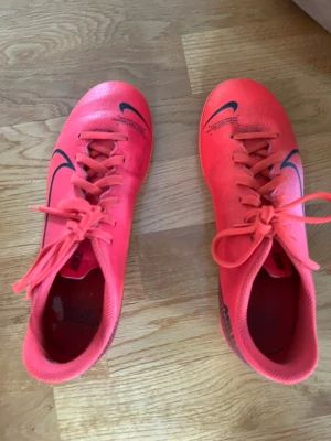 Nike Mercurial rosa fotbollsskor - Säljer ett par Nike Mercurial fotbollsskor i orange färg med svarta detaljer och Nike-logga. Skorna har snörning och dobbar för spel på gräs. Snygg, sportig design i syntetmaterial som ger bra grepp och känsla på planen.