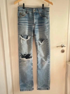 Ljusblå ripped jeans - Säljer ett par ljusblå straight fit jeans med stora slitningar och hål på båda benen. Jeansen har klassisk femficksdesign, hög midja och är tillverkade i denim. Perfekta för en avslappnad och trendig look.