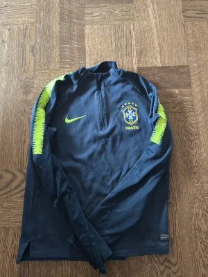 Nike Brasil långärmad träningströja strl: 137-147 - Mörkblå långärmad träningströja från Nike med halv dragkedja och Brasilien-emblem på bröstet. Neongula detaljer längs axlarna och Nike-logga på framsidan. Perfekt för fotbollsträning eller sportiga aktiviteter.