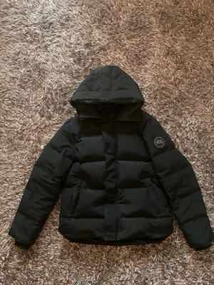 Svart pufferjacka från Canada Goose - Snygg svart pufferjacka från Canada Goose med huva och klassisk patch på ärmen. Jackan har dragkedja framtill, två sidofickor och en boxig passform. Perfekt för kalla dagar när du vill hålla dig varm och stilren.