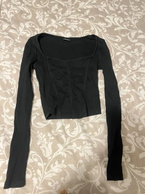 Svart ribbad topp från Gina Tricot - Trendig svart långärmad topp från Gina Tricot med ribbad struktur och markerade sömmar framtill. Toppen har en fyrkantig urringning och är croppad, vilket ger en snygg och modern look.