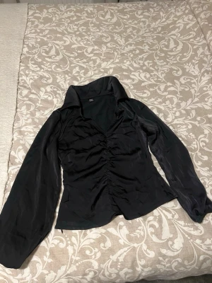 Svart blus med puffärm från SHEIN - Stilren svart blus från SHEIN med långa puffärmar och snygg rynkning framtill. Blusen har en djup krage och är tillverkad i ett glansigt, tunt material som ger en elegant känsla. Perfekt för att lyfta din outfit med en trendig touch.