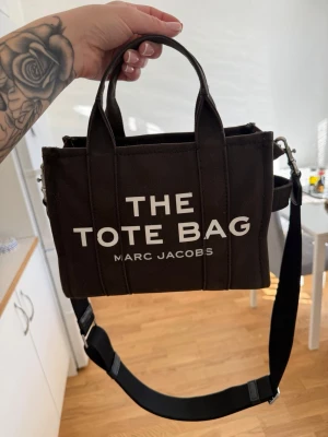 The Tote Bag från Marc Jacobs - Trendig svart handväska från Marc Jacobs med tydlig vit text 'THE TOTE BAG' på framsidan. Väskan är i canvasmaterial och har både handtag och justerbar axelrem för flexibel användning. Perfekt för dig som gillar stilrena och praktiska accessoarer. KVITTO FINNS!