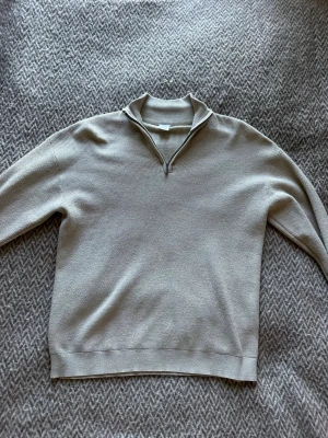Beige stickad halvzip från Lindex - Snygg beige stickad tröja från Lindex med halvzip och hög krage. Tröjan har ribbstickade muddar och är perfekt för lager-på-lager. Enkel och stilren design som passar till det mesta.