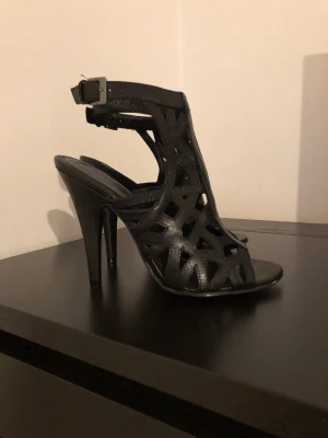 Svarta klackade pumps med utskurna detaljer - Snygga svarta pumps med hög klack och trendiga utskurna detaljer. Skorna har öppen tå, justerbart spänne vid ankeln och är tillverkade i äkta läder. Perfekta för dig som vill sticka ut med en edgy och modern look.