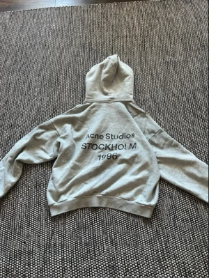 Grå hoodie från Acne Studios - Snygg grå hoodie från Acne Studios med tryck på ryggen där det står 'Acne Studios STOCKHOLM 1996'. Klassisk passform med huva och ribbade muddar. Perfekt för en chill och trendig look. Materialet känns mjukt och skönt mot huden.