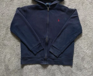 Mörkblå hoodie från Polo Ralph Lauren - Mörkblå hoodie med dragkedja från Polo Ralph Lauren. Klassisk design med huva, fickor framtill och den ikoniska röda logotypen broderad på bröstet. Tillverkad i mjukt bomullsmaterial som känns skönt mot huden. Perfekt för en avslappnad och stilren look.