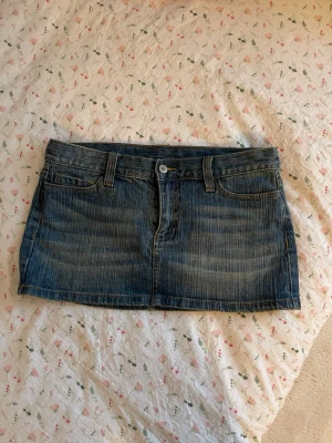 Blå jeanskjol i kort modell - Säljer en klassisk blå jeanskjol i kort modell med fem fickor och bälteshällor. Kjolen har en knapp och dragkedja framtill samt snygga sömmar och slitningar för en cool look. Perfekt till sneakers eller boots!