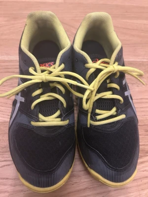 Svarta Asics Gel-Tactic sneakers - Säljer ett par svarta Asics Gel-Tactic sneakers med gula detaljer och snören. Skorna har meshmaterial på ovansidan för bra ventilation och en låg profil. Perfekta för dig som vill ha bekväma och sportiga sneakers med cool design.