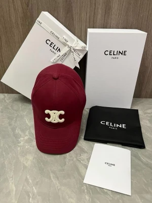 Vinröd keps från Celine med logga - Snygg vinröd keps från Celine med den ikoniska vita Triomphe-loggan broderad framtill. Klassisk böjd skärm och justerbar rem bak. Tillverkad i bomullstyg som ger en skön och stilren look. Perfekt accessoar för att lyfta din outfit.