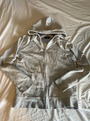 Ljusgrå zip hoodie från GANT - Snygg ljusgrå hoodie från GANT med dragkedja framtill och klassisk huva. Tröjan har två fickor på magen och GANT-logga broderad på bröstet. Tillverkad i mjukt bomullsmaterial som känns skönt mot huden. Perfekt för en avslappnad och stilren look.
