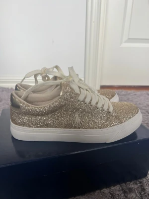 Glittriga guldiga sneakers från Polo - Snygga sneakers från Polo Ralph Lauren i glittrigt guld med vita snören och vit sula. Låg modell med klassisk Polo-logga på sidan. Perfekta för dig som vill sticka ut med en trendig och glamorös look. Materialet är syntet med glitterfinish. Skorna är helt nya och aldrig använda, har kvar boxen och kvittot, skorna är i storlek 35 men passar ändå mig som har storlek 36/37